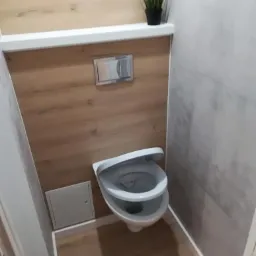 Nowoczesna toaleta podwieszana z drewnopodobnymi i szarymi płytkami, ozdobiona rośliną w czarnej doniczce na półce.