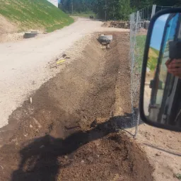 Wykopany rów w ziemi z widocznymi betonowymi studzienkami, narzędziami i paletami w tle, sfotografowany z perspektywy kierowcy pojazdu z widocznym lusterkiem bocznym.