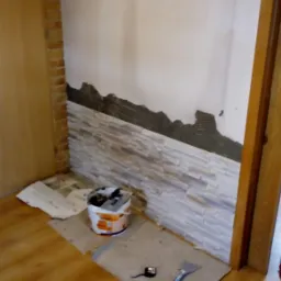 Wnętrze w trakcie prac: fragment ściany z częściowo ułożoną dekoracyjną okładziną kamienną. Widoczne narzędzia i materiały budowlane na podłodze.