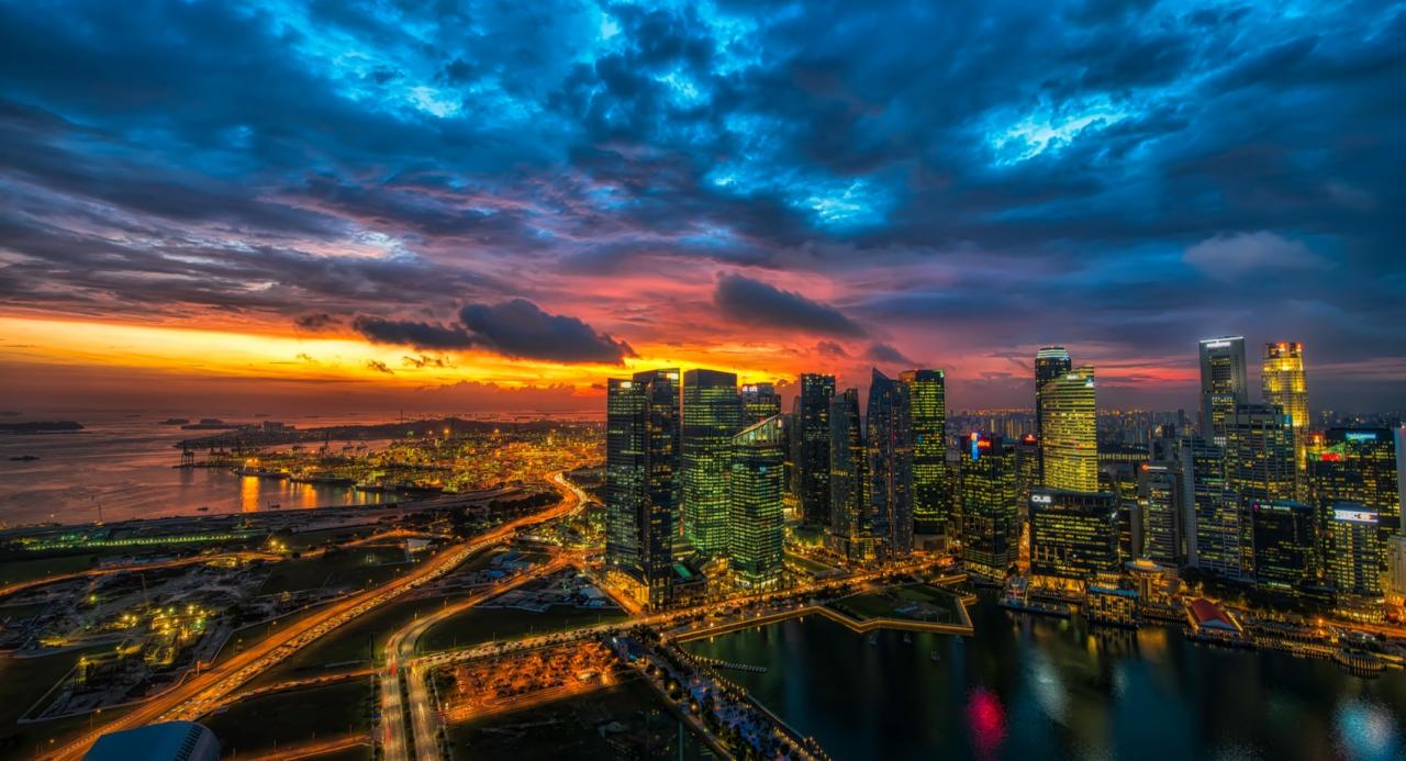 Panorama miasta nocą z drapaczami chmur oświetlonymi ciepłym światłem, pod dramatycznym, wielobarwnym niebem o zachodzie słońca.