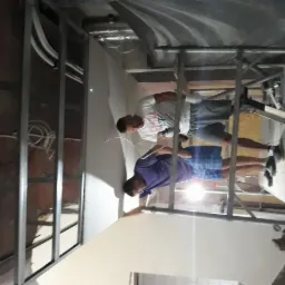 Dwaj mężczyźni pracują na rusztowaniu przy montażu metalowej konstrukcji pod płytę karton gips na ścianie. Widoczne przewody elektryczne i elementy instalacji. Ujęcie z dołu, w trakcie prac...