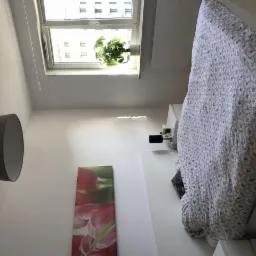 Jasne wnętrze sypialni z ł&oacute;żkiem w kwiatowy wz&oacute;r, lampą z abażurem i obrazem tulipan&oacute;w na ścianie, roślina w doniczce na parapecie okna.