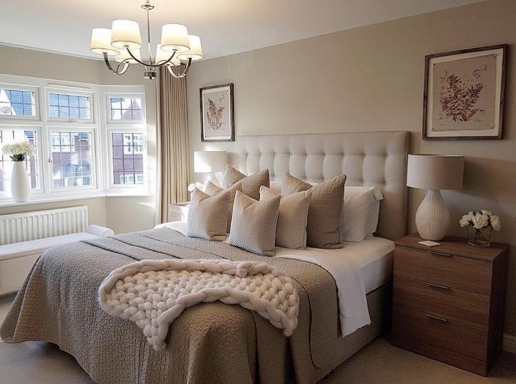 Elegancka sypialnia w odcieniach beżu z dużym łóżkiem, ozdobną narzutą z grubego splotu, poduszkami w różnych teksturach, oświetlona naturalnym światłem z okna wykuszowego i lampami nocnymi.
