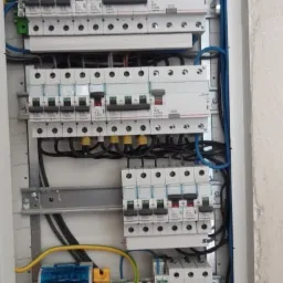 Rozdzielnia elektryczna w trakcie montażu, widoczne zabezpieczenia różnicowoprądowe, bezpieczniki oraz kable w kolorach niebieskim, żółto-zielonym i czarnym, umieszczone w białej obudowie.