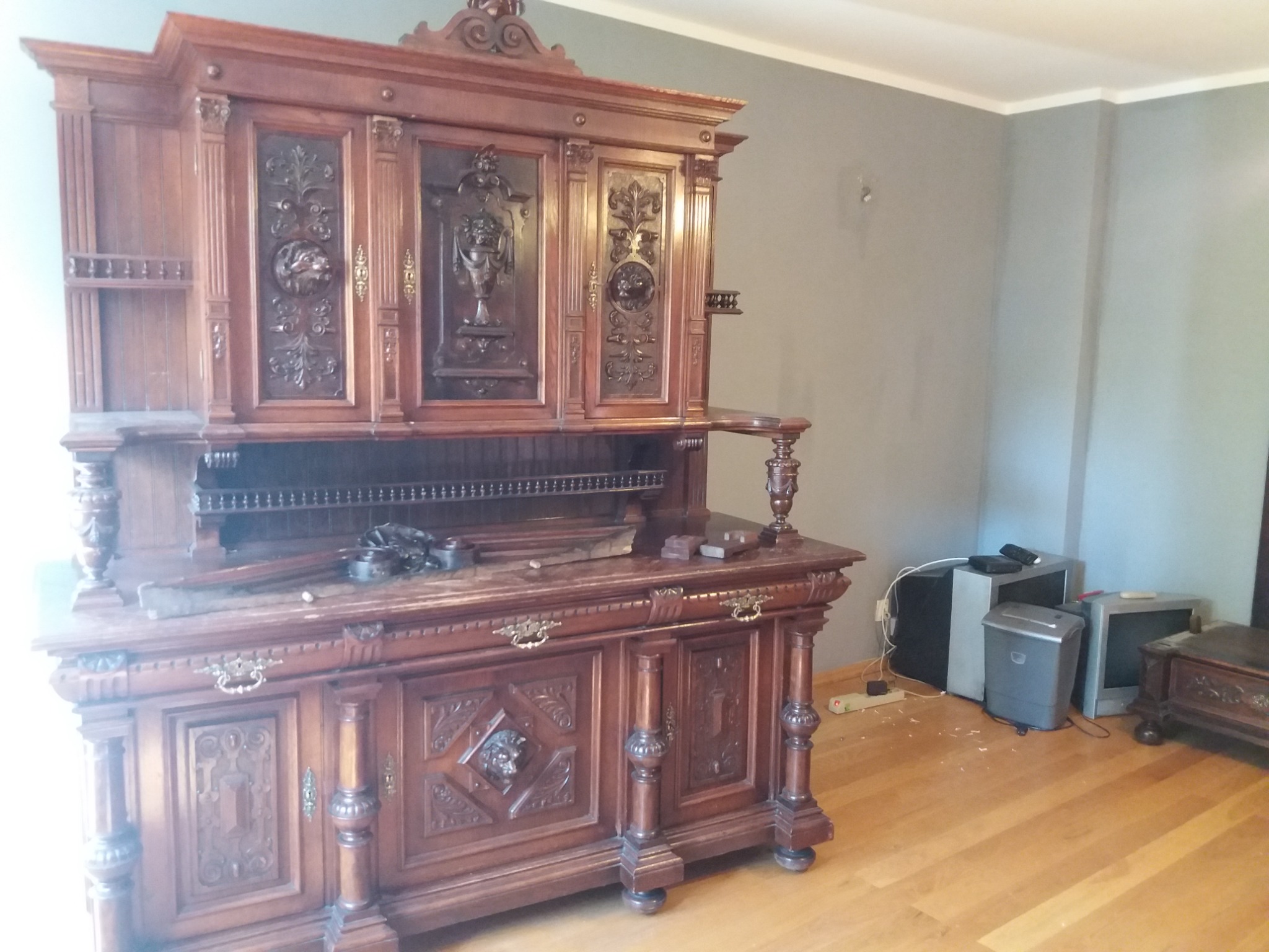 Antyczny, rzeźbiony kredens z ciemnego drewna, widoczny podczas transportu, z akcesoriami do pakowania na blacie i sprzętem RTV w tle.