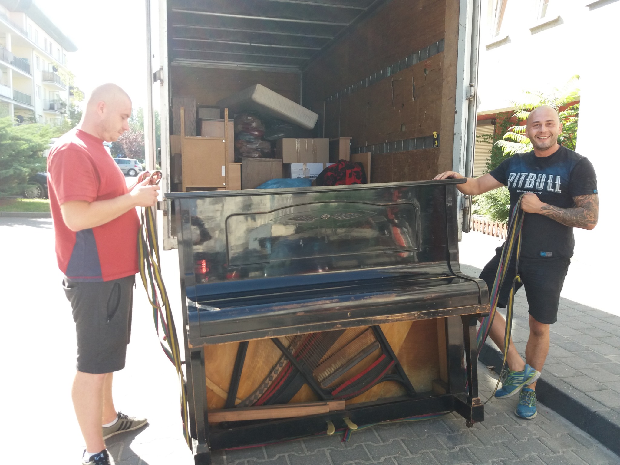 Dwóch mężczyzn transportuje pianino do wnętrza ciężarówki; w tle widoczne kartony i materac.