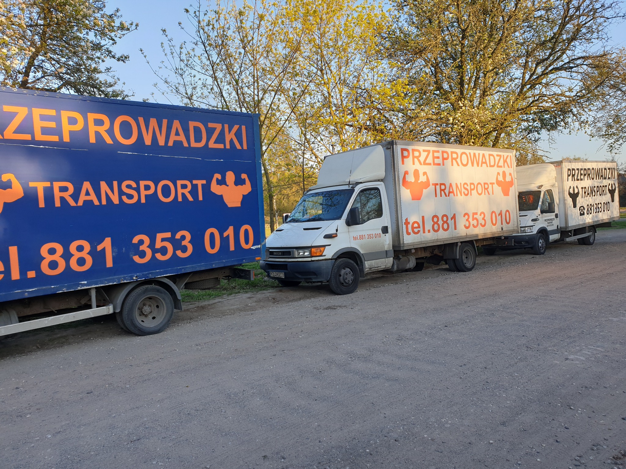 Flota trzech samochodów dostawczych z pomarańczowym logo firmy transportowej i numerem telefonu, zaparkowana na żwirowej drodze w słoneczny dzień.