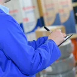 Osoba w niebieskim uniformie sporządza notatki na schowku, w tle widoczne materiały budowlane i magazynowe.