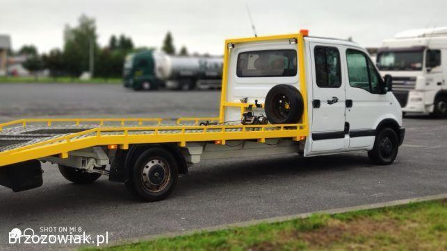 Biała laweta z żółtą platformą na parkingu, z zapasowym kołem i wciągarką na platformie, w tle widoczne inne pojazdy ciężarowe.