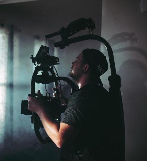 Operator filmowy z brodą, w czapce z napisem, obsługuje profesjonalny stabilizator kamery podczas nagrywania w pomieszczeniu.