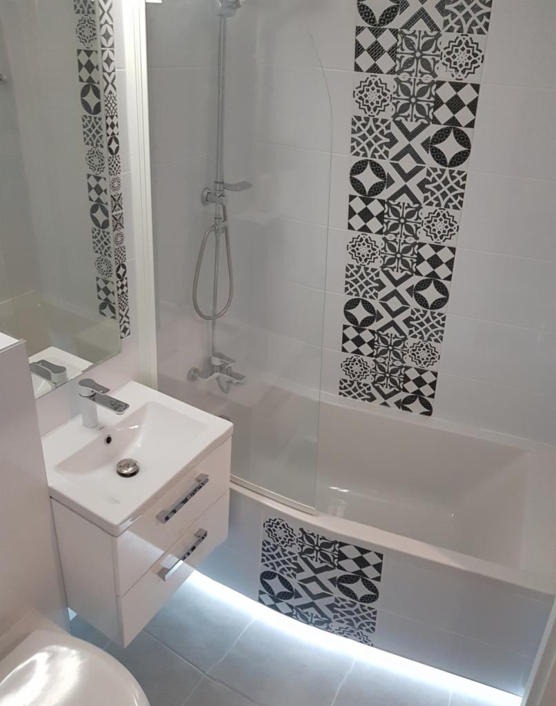 Nowoczesna łazienka z białą szafką pod umywalką, chromowaną armaturą i wanną z parawanem, akcentowana czarno-białymi płytkami dekoracyjnymi i subtelnym oświetleniem LED pod szafką.