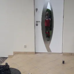 Nowoczesne białe drzwi wejściowe z dekoracyjnym, srebrnym elementem w kształcie łzy, widoczne ślady montażu: puszka pianki montażowej i worek na śmieci na kafelkowej podłodze.