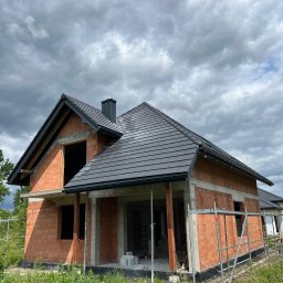 Usługi budowlane Piotr Przybytek - Surowy dom z czerwonej cegły pod ciemnym, zachmurzonym niebem. Widoczny dach z czarnej dachówki, brak okien i drzwi. Etap budowy, rusztowania.