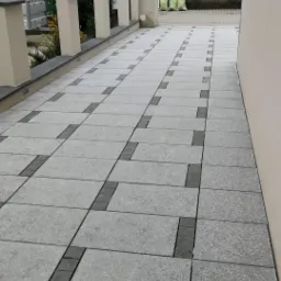 Ułożona kostka brukowa w odcieniach szarości tworząca wzór na podjeździe, widoczne kolumny ogrodzeniowe i fragment budynku w tle.