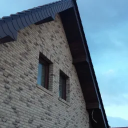 Dom z cegły klinkierowej z ciemną dachówką i widocznym kominem, sfotografowany pod kątem w pochmurny dzień.