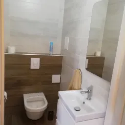 Nowoczesna łazienka z szarymi płytkami ściennymi, drewnopodobną zabudową, podwieszaną toaletą, białą umywalką z szafką i lustrem bez ramy.