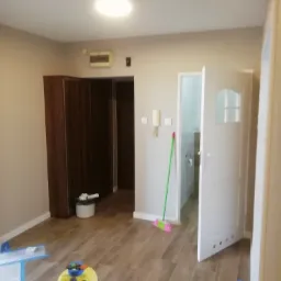 Wnętrze pokoju po remoncie z panelami podłogowymi, widoczny odkurzacz, szafa w zabudowie, otwarta biała łazienkowa, ściany pomalowane na jasny beż.