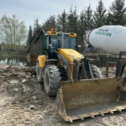 Żółto-czarna koparko-ładowarka Mecalac nabiera ziemię w pobliżu zbiornika wodnego; w tle betonomieszarka Drogmost Beton i las.