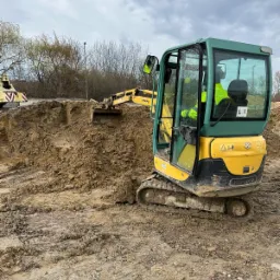 Żółto-zielona mini koparka Yanmar w trakcie prac ziemnych, operator w odblaskowej kamizelce, teren budowy z błotnistym podłożem i wykopaną ziemią.