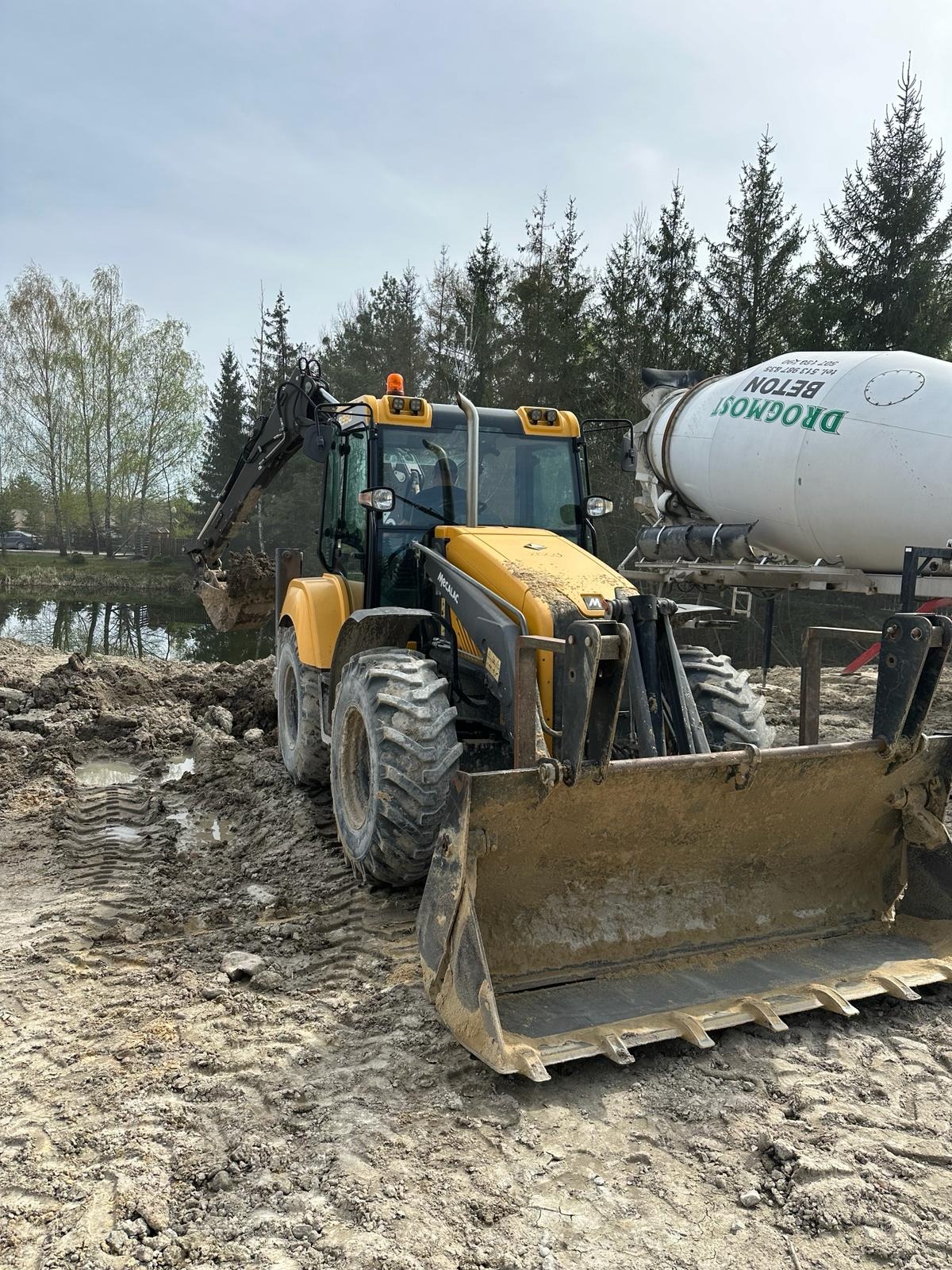 Żółto-czarna koparko-ładowarka Mecalac nabiera ziemię w pobliżu zbiornika wodnego; w tle betonomieszarka Drogmost Beton i las.