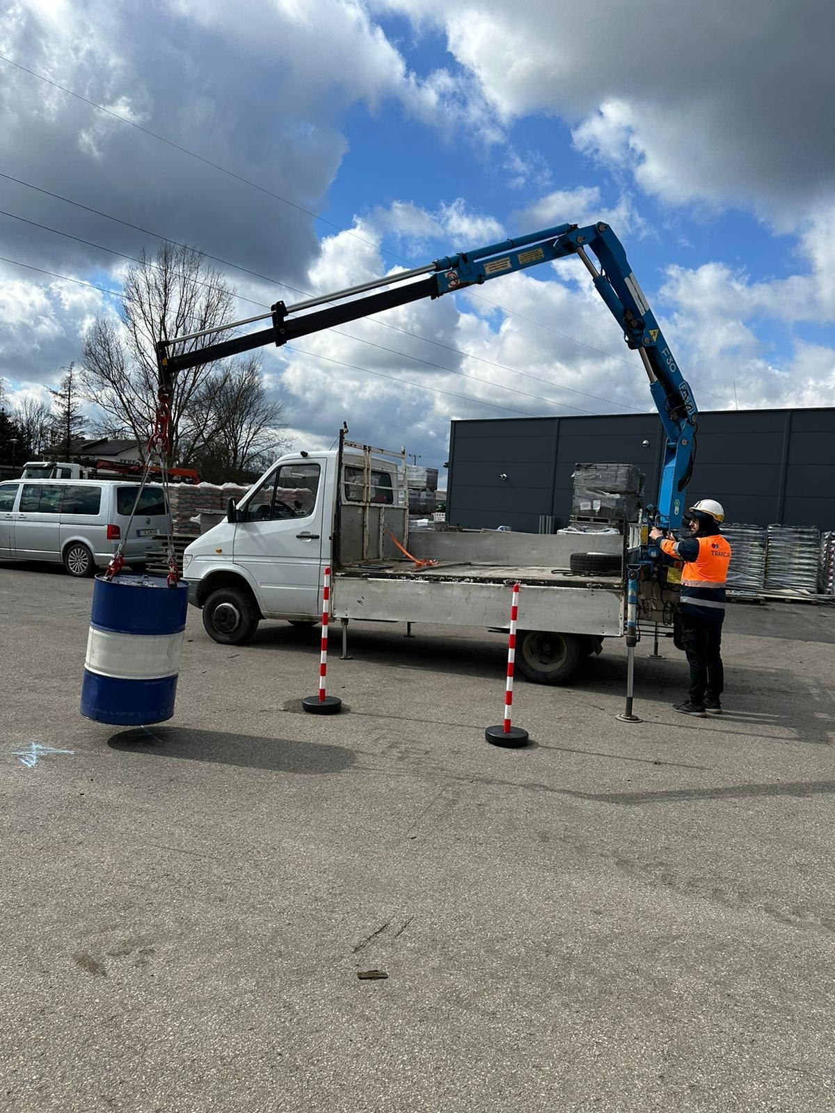 Pracownik w kamizelce i kasku obsługuje niebieski żuraw zamontowany na białej ciężarówce, podnoszący niebiesko-białą beczkę na placu budowy, z widocznymi słupkami ostrzegawczymi.