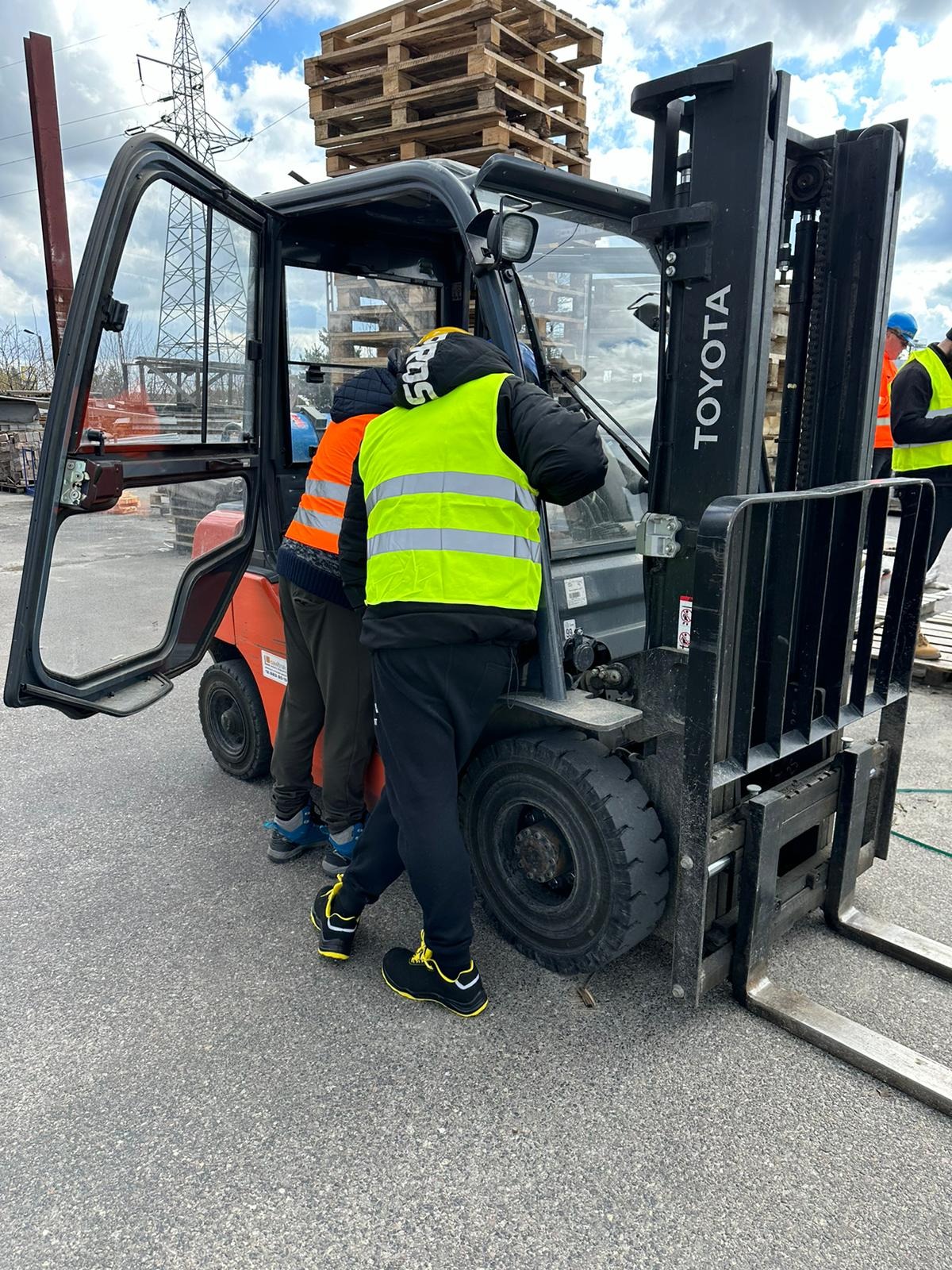 Dwóch mężczyzn w odblaskowych kamizelkach przy otwartej kabinie pomarańczowego wózka widłowego Toyota, palety w tle