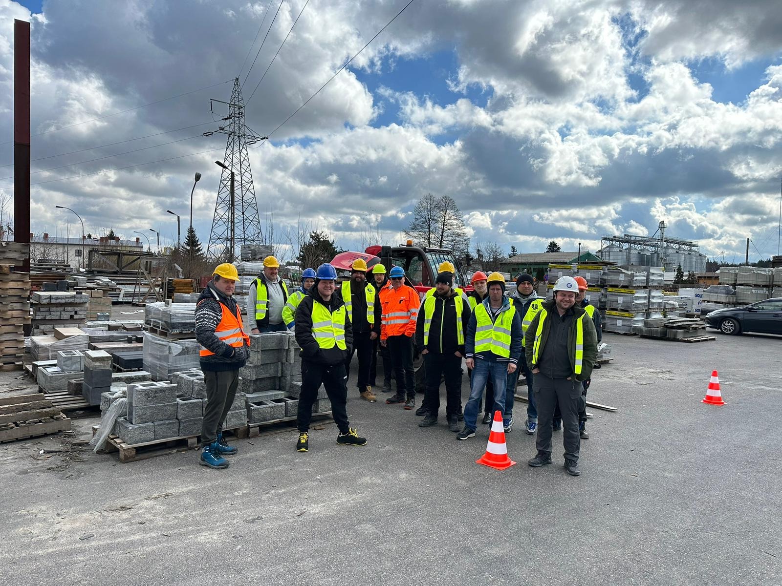 Grupa pracowników budowlanych w kaskach i kamizelkach odblaskowych pozuje na tle składu materiałów budowlanych, widoczne palety z kostką brukową, słup wysokiego napięcia i zachmurzone niebo.