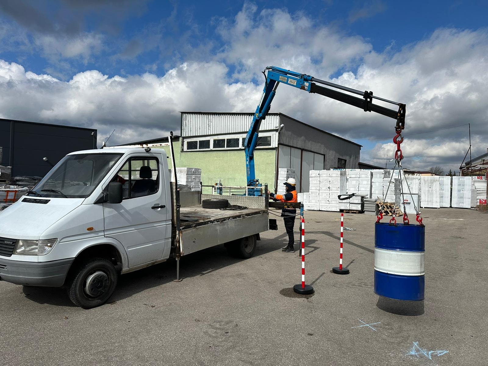 Pracownik w kasku i kamizelce obsługuje ramię dźwigu zamontowanego na białym samochodzie dostawczym, podnosząc niebiesko-białą beczkę na tle magazynu i składowanych materiałów budowlanych.