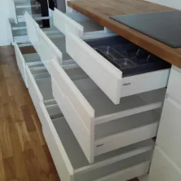 Białe szuflady kuchenne z systemem Blum, wysunięte, z drewnianym blatem i płytą indukcyjną w tle, widoczna wkładka na sztućce.