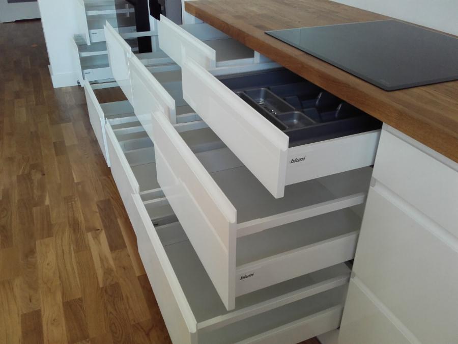 Białe szuflady kuchenne z systemem Blum, wysunięte, z drewnianym blatem i płytą indukcyjną w tle, widoczna wkładka na sztućce.