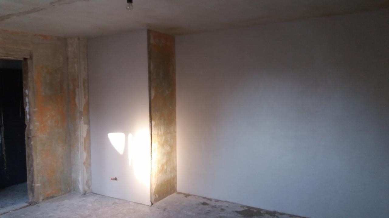 Wnętrze pokoju w trakcie prac wykończeniowych, widoczna ściana po nałożeniu gładzi i wstępnym szlifowaniu, surowy beton na podłodze, niezabezpieczone narożniki, oraz widoczny kabel elektryczny...