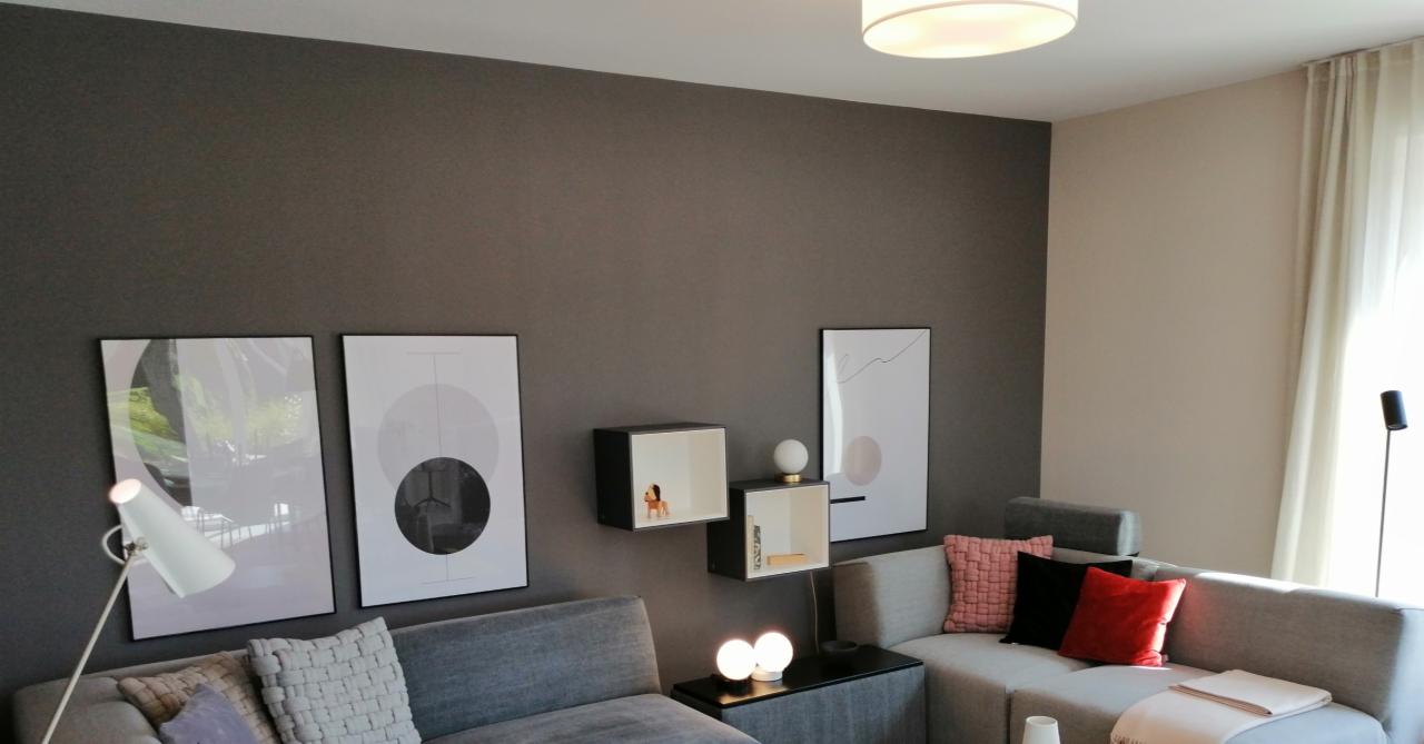 Nowoczesny salon z szarą ścianą, na której wiszą trzy grafiki w czarnych ramkach i dwie czarno-białe kostki pełniące funkcję półek, szary narożnik z kolorowymi poduszkami, oświetlony lampą sufitową...