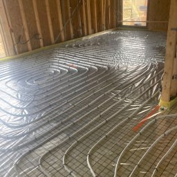 Lukasz Dyrek Łuk-komplex - Wnętrze domu szkieletowego w trakcie budowy, widoczne rury ogrzewania podłogowego ułożone na siatce zbrojeniowej, przytwierdzone pomarańczowymi taśmami, ściany z drewnianych belek.