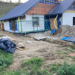 Lukasz Dyrek Łuk-komplex - Wznoszony dom jednorodzinny z ciemnoszarą dachówką, częściowo ocieplony, z widoczną konstrukcją drewnianą i rusztowaniem, na działce z ziemią, gruzem i workami na śmieci.