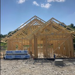 Lukasz Dyrek Łuk-komplex - Konstrukcja szkieletowa domu w trakcie budowy, widoczne drewniane elementy nośne ścian i dachu, na pierwszym planie paczki materiałów budowlanych z logo producenta, tło stanowi błękitne niebo...