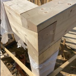 Lukasz Dyrek Łuk-komplex - Połączenie belek z jasnego drewna w konstrukcji szkieletowej domu, częściowo zabezpieczonych folią, widoczny fragment pracującego dekarza.