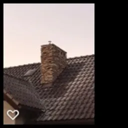 Ciemnobrązowy dach z dachówki ceramicznej z kominem obłożonym brązowym kamieniem, widziany z dołu pod lekkim kątem, na tle jasnego nieba o zachodzie słońca.