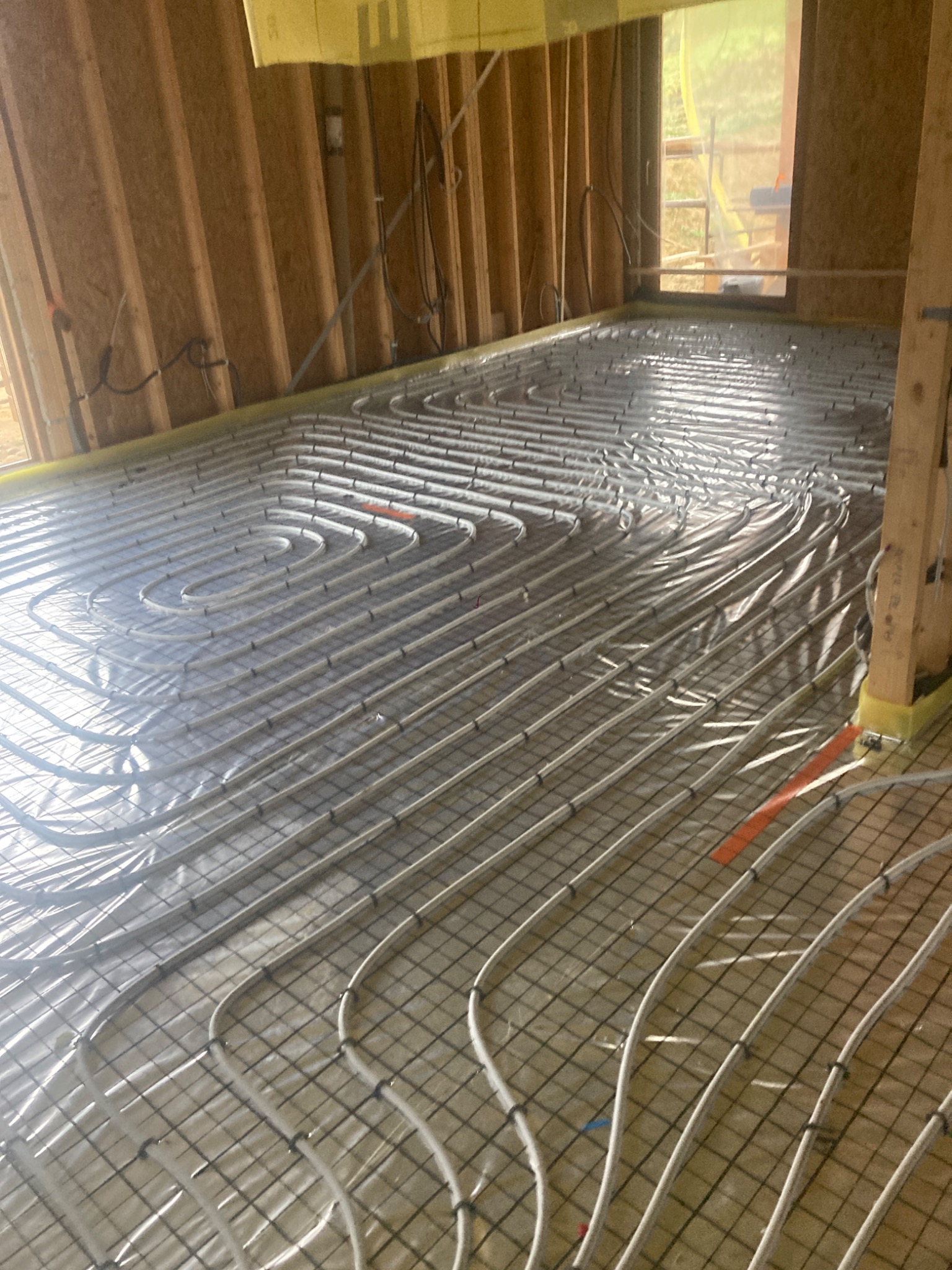 Wnętrze domu szkieletowego w trakcie budowy, widoczne rury ogrzewania podłogowego ułożone na siatce zbrojeniowej, przytwierdzone pomarańczowymi taśmami, ściany z drewnianych belek.