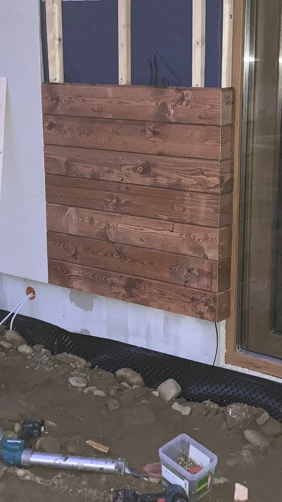 Montaż drewnianej elewacji na ścianie budynku, widoczne narzędzia i materiały budowlane na ziemi.