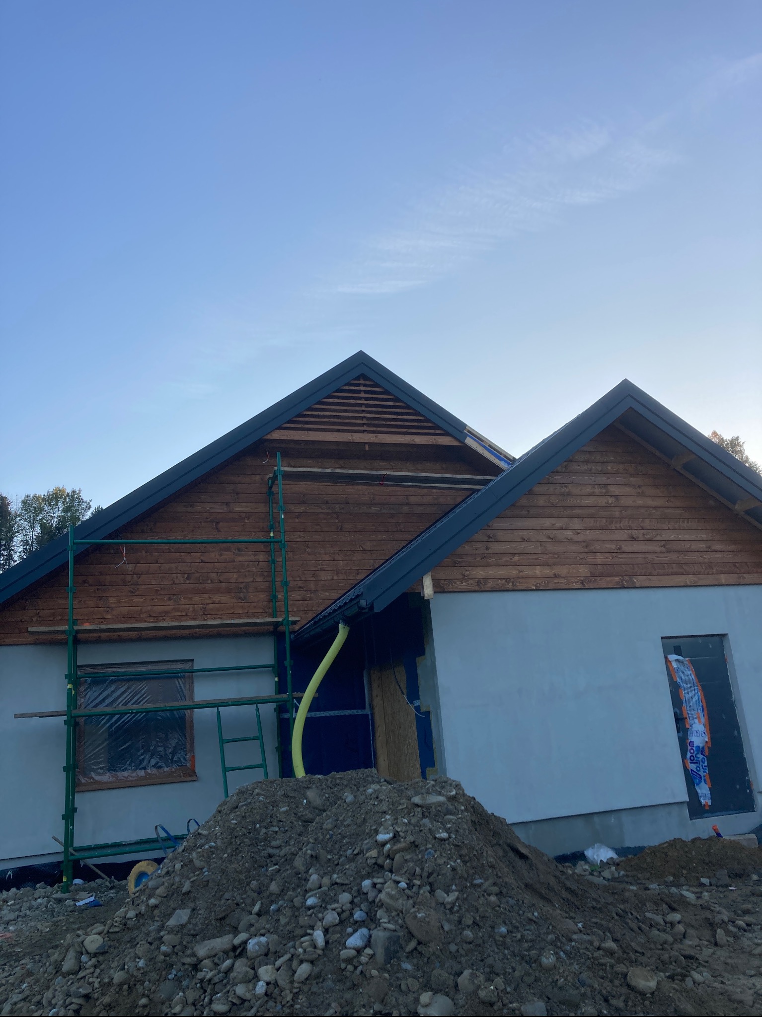 Dom w trakcie budowy z drewnianą elewacją szczytową, zielonymi rusztowaniami i stertą ziemi przed budynkiem w słoneczny dzień.