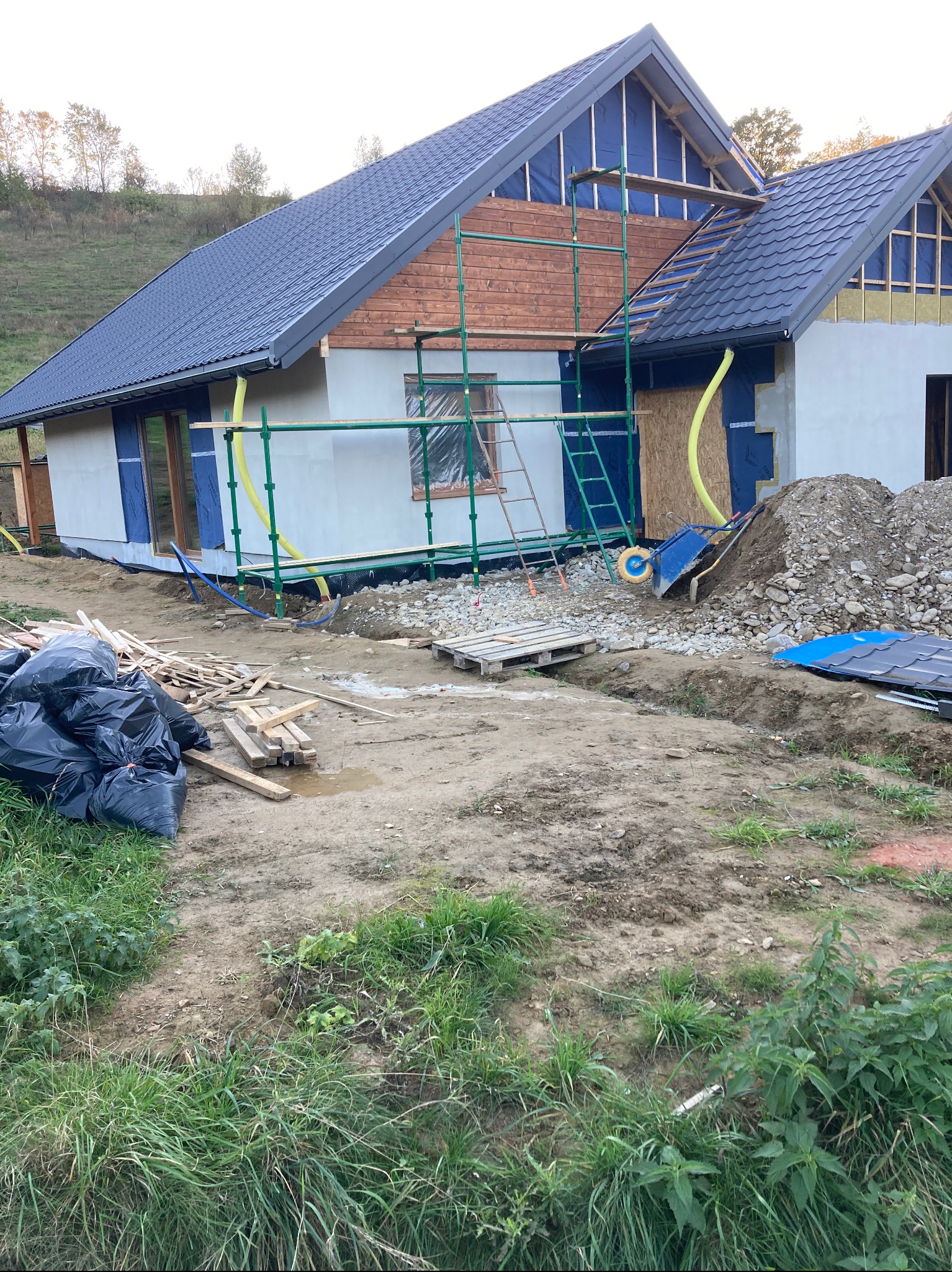 Wznoszony dom jednorodzinny z ciemnoszarą dachówką, częściowo ocieplony, z widoczną konstrukcją drewnianą i rusztowaniem, na działce z ziemią, gruzem i workami na śmieci.
