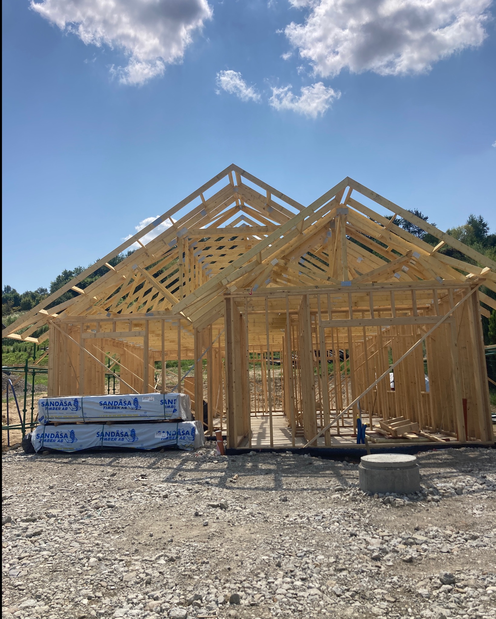 Konstrukcja szkieletowa domu w trakcie budowy, widoczne drewniane elementy nośne ścian i dachu, na pierwszym planie paczki materiałów budowlanych z logo producenta, tło stanowi błękitne niebo...