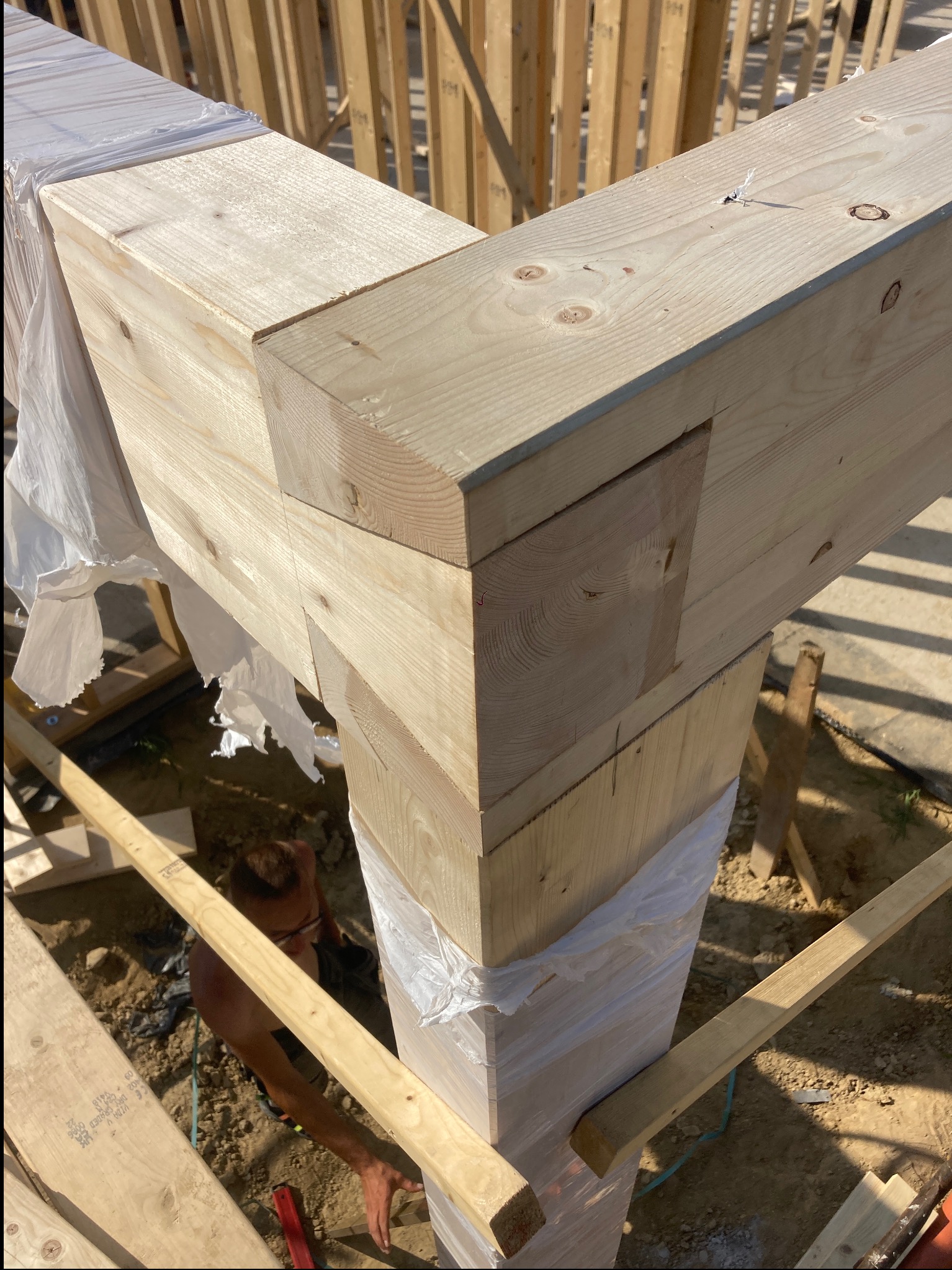 Połączenie belek z jasnego drewna w konstrukcji szkieletowej domu, częściowo zabezpieczonych folią, widoczny fragment pracującego dekarza.