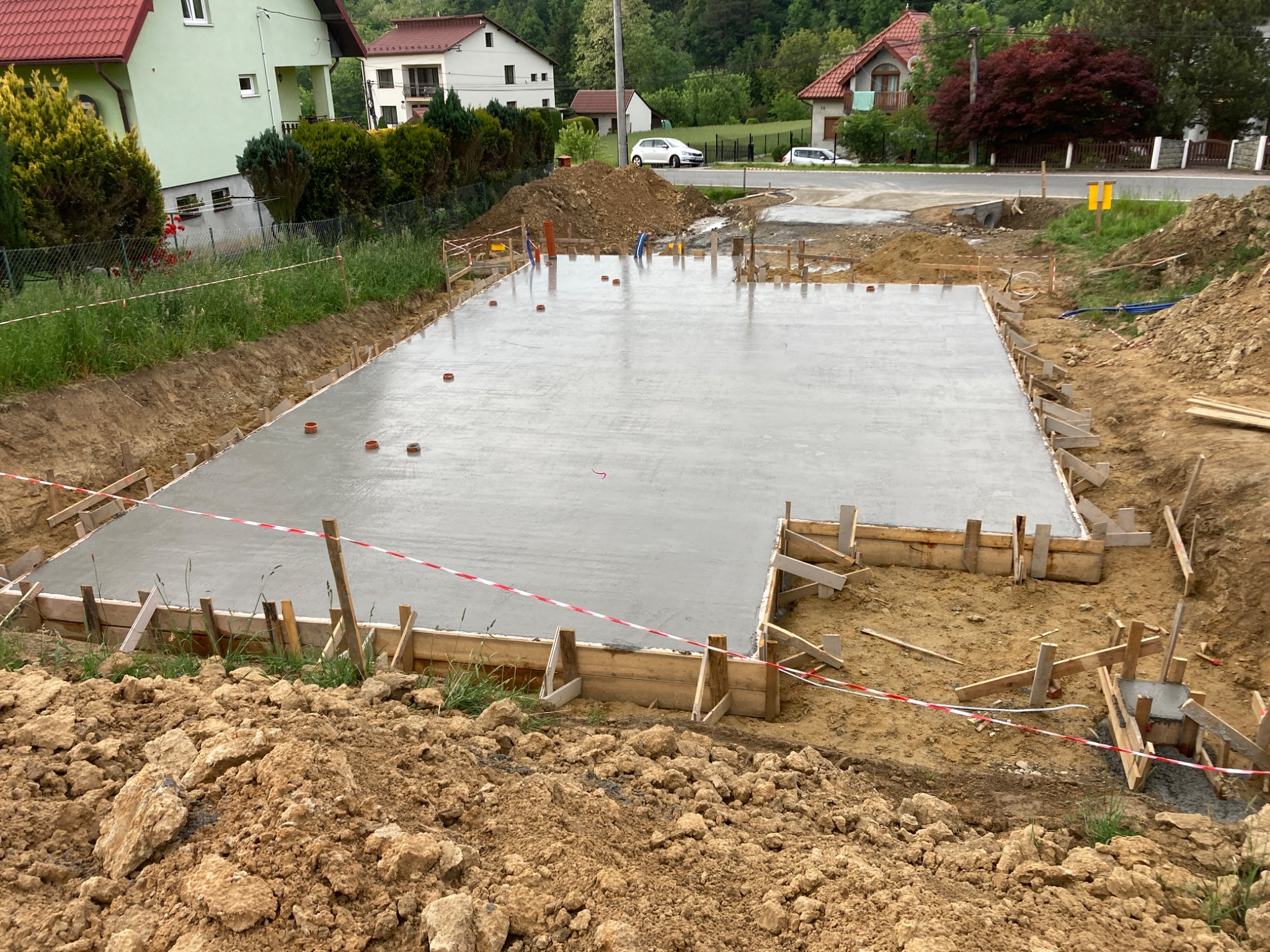 Wylany fundament pod budowę domu jednorodzinnego, widoczne szalunki i poziomowanie, w tle zabudowa mieszkalna i droga.