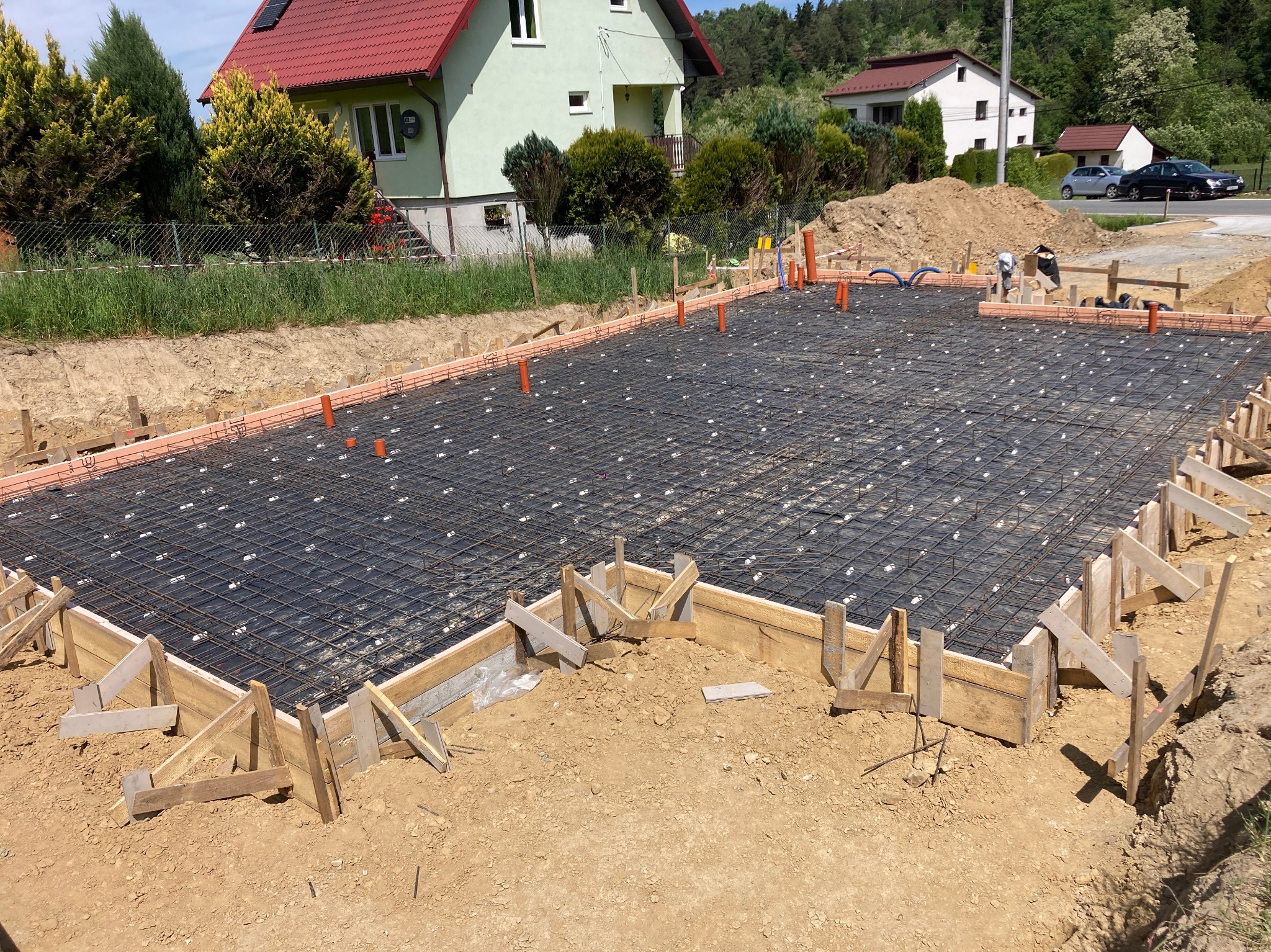 Przygotowanie podłoża pod wylewkę fundamentową: zbrojenie siatką stalową, szalunek z desek z drewnianymi wspornikami i wystające rury instalacyjne, na tle domów jednorodzinnych.