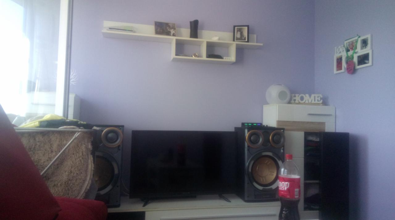 Telewizor na białej szafce RTV, obok kolumny głośnikowe i butelka coli, na ścianie półka z dekoracjami, w tle fioletowa ściana.