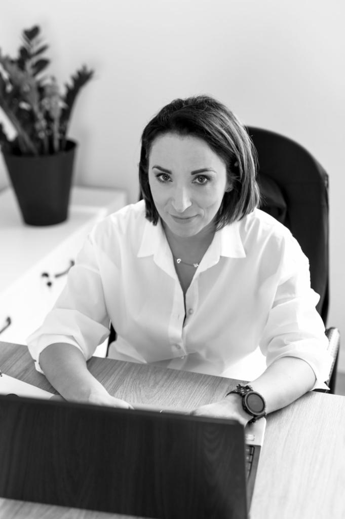 Portret kobiety w białej koszuli siedzącej za biurkiem z laptopem, z zegarkiem na nadgarstku, w tle roślina doniczkowa i białe meble, zdjęcie czarno-białe.