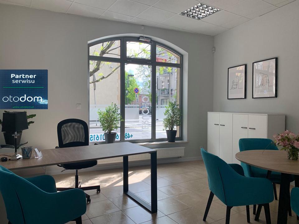 Jasne biuro agencji nieruchomości z dużym oknem łukowym, biurkami, turkusowymi krzesłami i logo Partner serwisu Otodom na ścianie.
