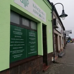 Zewnętrzny widok na biuro Kancelarii Adwokackiej z zielonymi ścianami, szyldem i oknem z wypisanymi usługami prawnymi, obok ulicy z przejeżdżającym samochodem.
