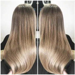 Długie, proste włosy z ombre, przechodzące od ciemnego blondu u nasady do jasnego blondu na końcach, widoczne z tyłu, z delikatnym połyskiem.